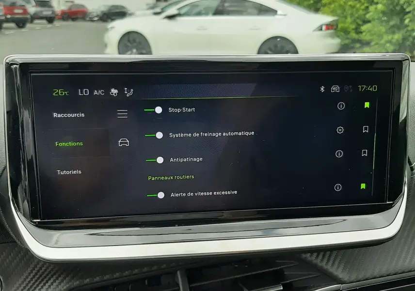 Vue intérieure centrée sur l'écran tactile du tableau de bord, avec en arrière-plan une Peugeot 208 gris Artense vue de profil côté gauche.