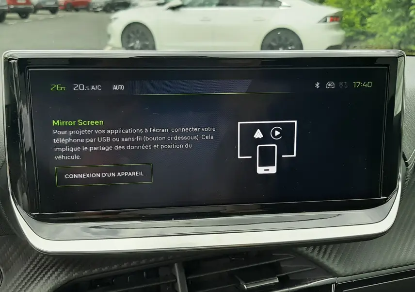 Écran tactile central du tableau de bord d’une Peugeot 208 1.2 100CH GT, avec vue floue extérieure sur une voiture blanche.