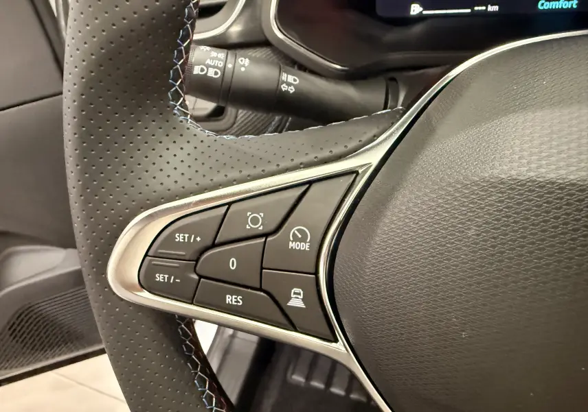 Gros plan sur les commandes gauche du volant perforé du Renault Captur blanc nacré, avec surpiqûres bleues et affichage numérique partiel.