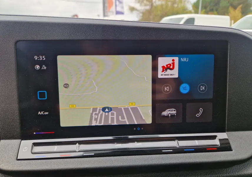 Écran tactile central du Volkswagen Caddy Cargo 2023 affichant la navigation et la radio NRJ, avec commandes climatisation en dessous.
