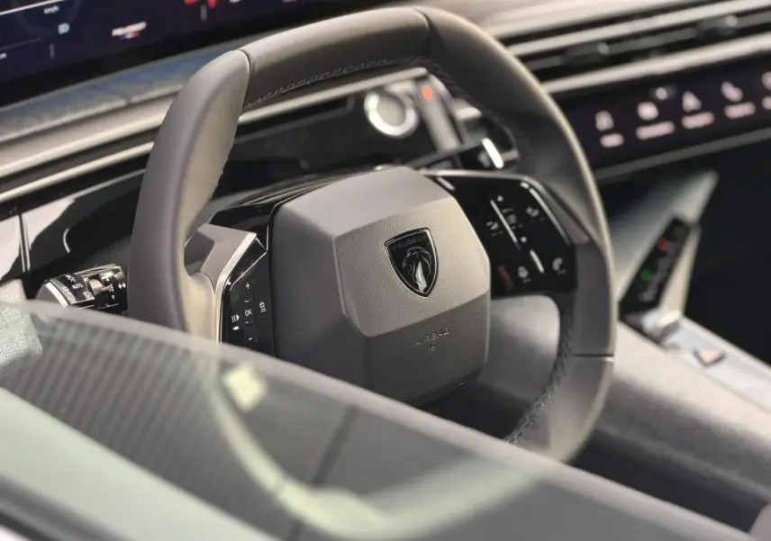 Gros plan sur le volant noir du Peugeot 3008 Hybrid 2025 avec logo lion et tableau de bord numérique en arrière-plan.