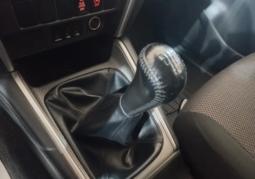 Gros plan sur le levier de vitesses manuel en cuir noir du Mitsubishi L200, avec console argentée et siège tissu gris visible.