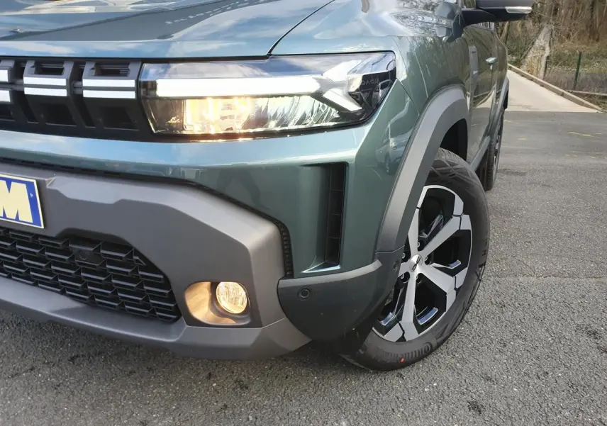 Gros plan sur l'avant droit du Dacia Duster vert cèdre, mettant en valeur le phare allumé et la jante alliage 18 pouces.