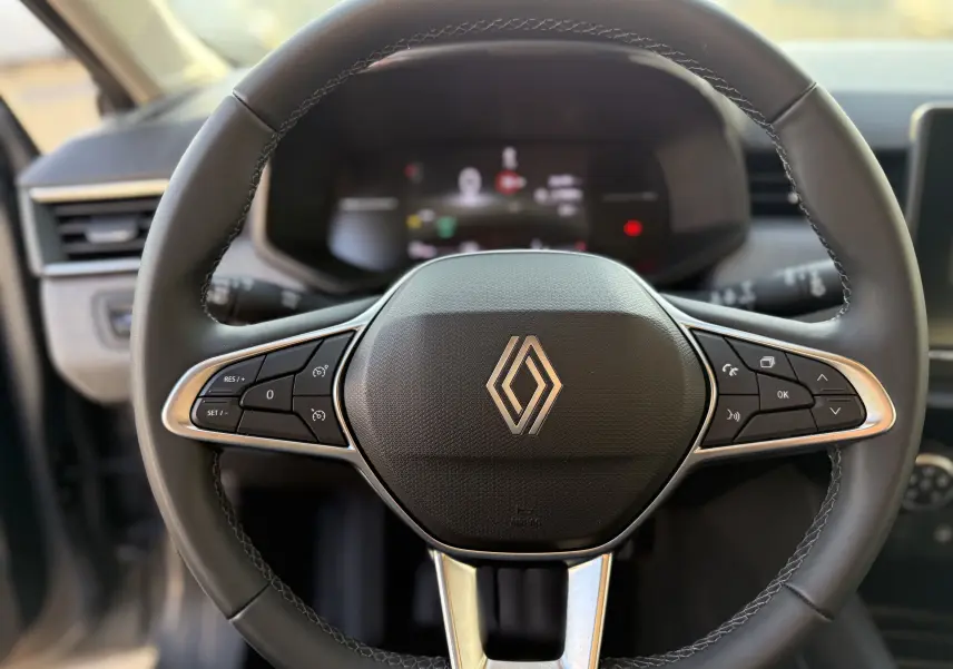 Gros plan sur le volant cuir noir de la Renault Clio V 2025 avec commandes intégrées et logo central argenté.
