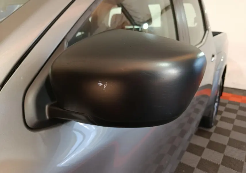 Gros plan sur le rétroviseur noir côté conducteur du Nissan Navara gris clair, vue partielle du profil avant.