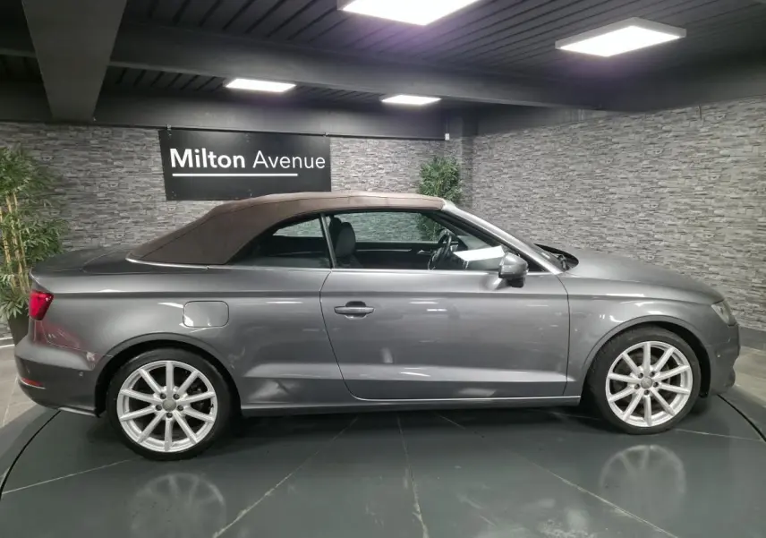 Audi A3 Cabriolet gris foncé vue de profil côté gauche avec capote tissu noire en intérieur showroom.