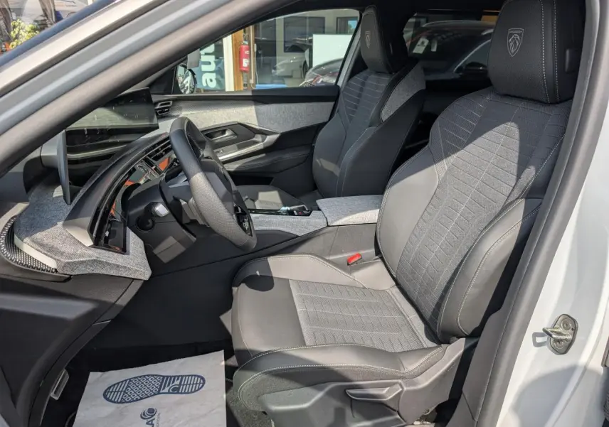 Vue intérieure côté conducteur du Peugeot 5008 blanc 2025, sièges tissu gris avec logo Peugeot brodé sur appuie-tête.