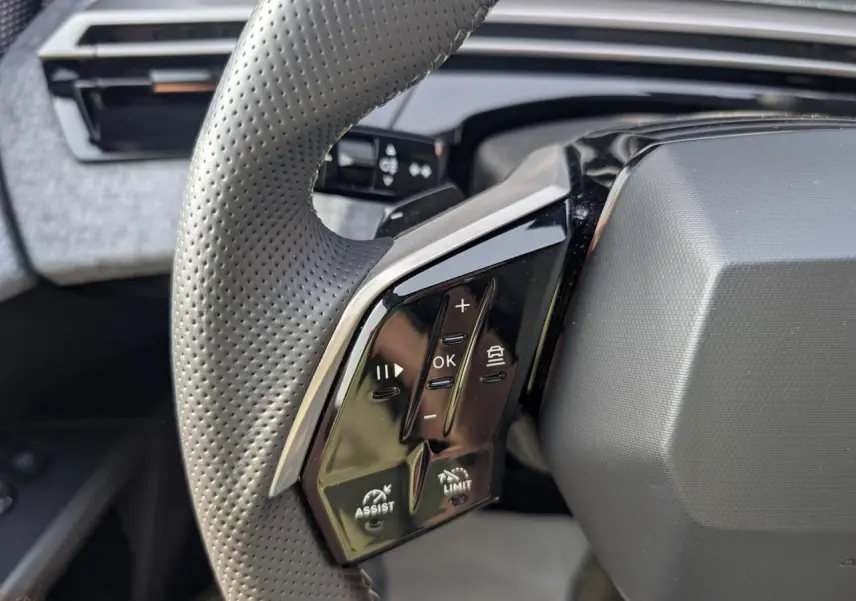 Gros plan sur les commandes gauche du volant perforé noir du Peugeot 5008 Hybrid 2025, avec boutons de régulateur et assistances.