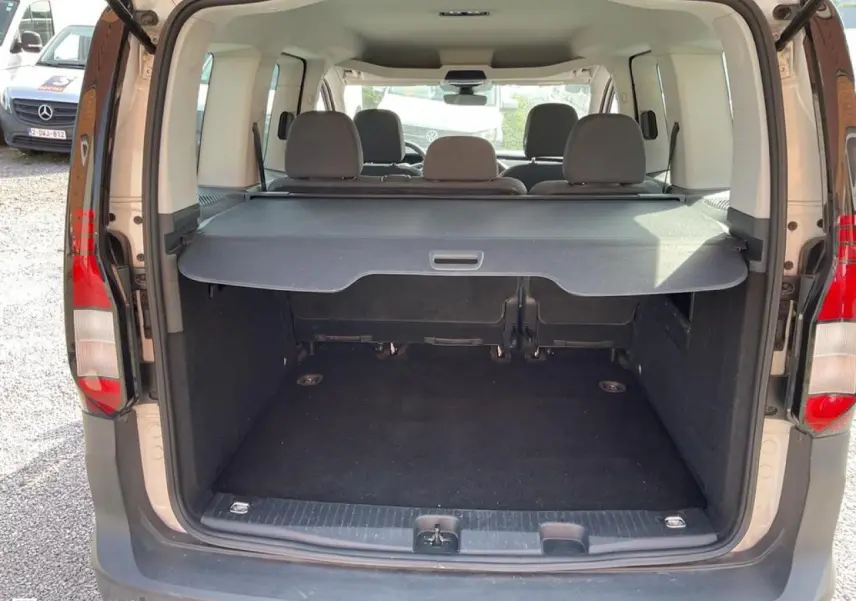 Vue arrière du coffre ouvert d'un Volkswagen Caddy blanc Candy 2024 avec cache-coffre enroulable et sièges arrière visibles.