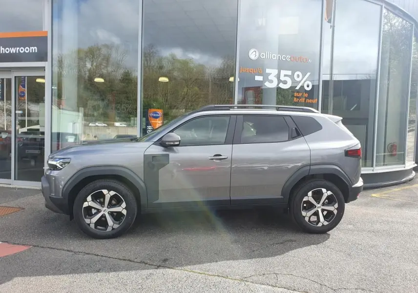 Vue de profil côté gauche d'un SUV Dacia gris schiste avec jantes alliage et barres de toit devant un showroom.