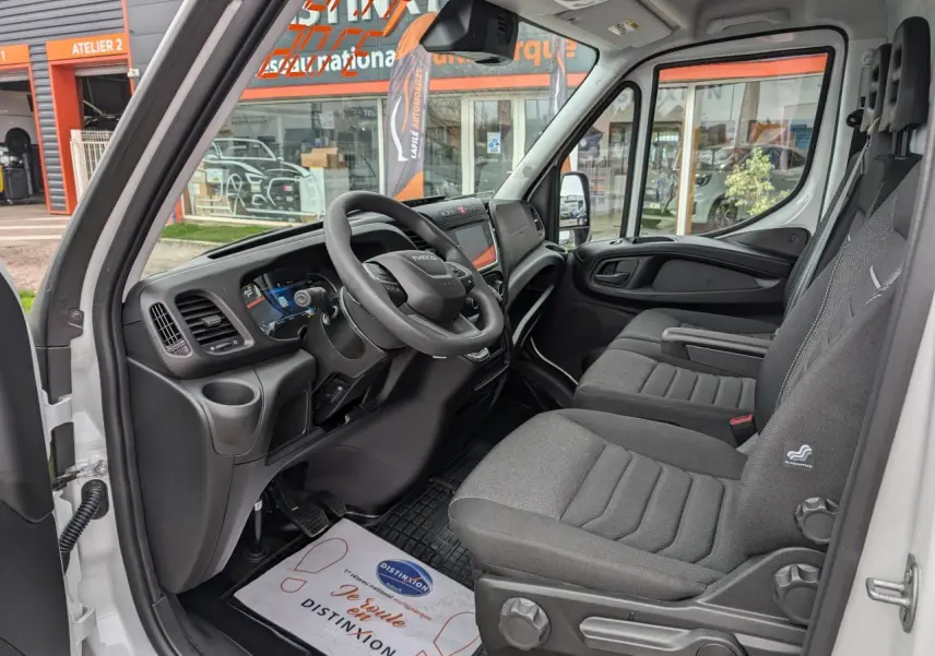 Vue intérieure côté conducteur de l'IVECO Daily blanc 2025, montrant tableau de bord moderne et sièges en tissu gris.