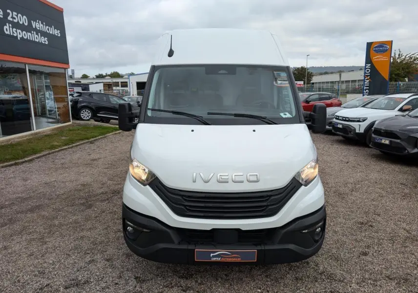 Fourgon Iveco Daily blanc vu de face, avec calandre noire et phares allumés sur un parking extérieur.