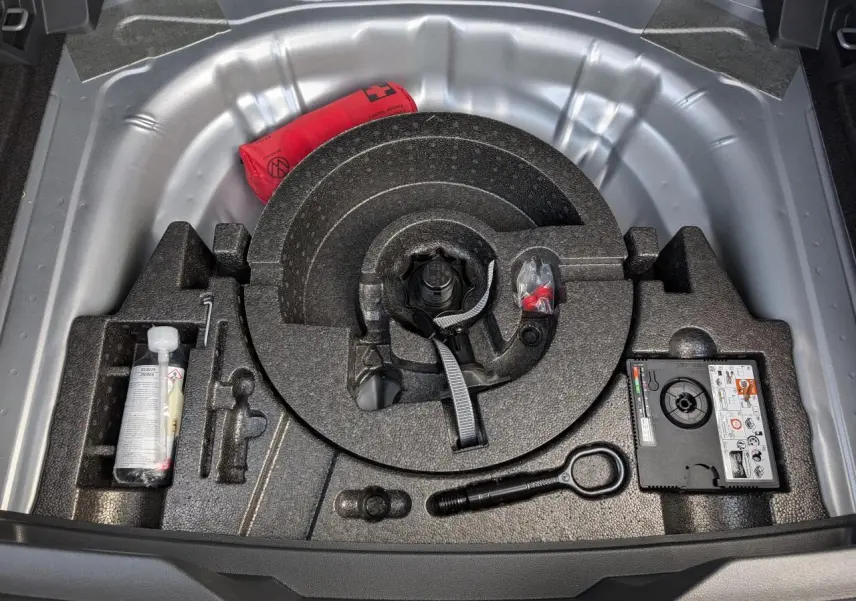 Vue plongeante sur le coffre ouvert d’un Volkswagen T-Roc gris, montrant la roue de secours et les outils de bord.