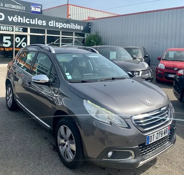 Peugeot 2008 gris métallisé vue 3/4 avant droit, avec calandre chromée et barres de toit visibles sur parking.