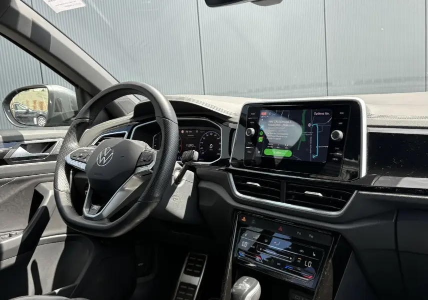 Vue intérieure côté conducteur du Volkswagen T-Roc gris Indium, avec volant multifonction et écran tactile central allumé.