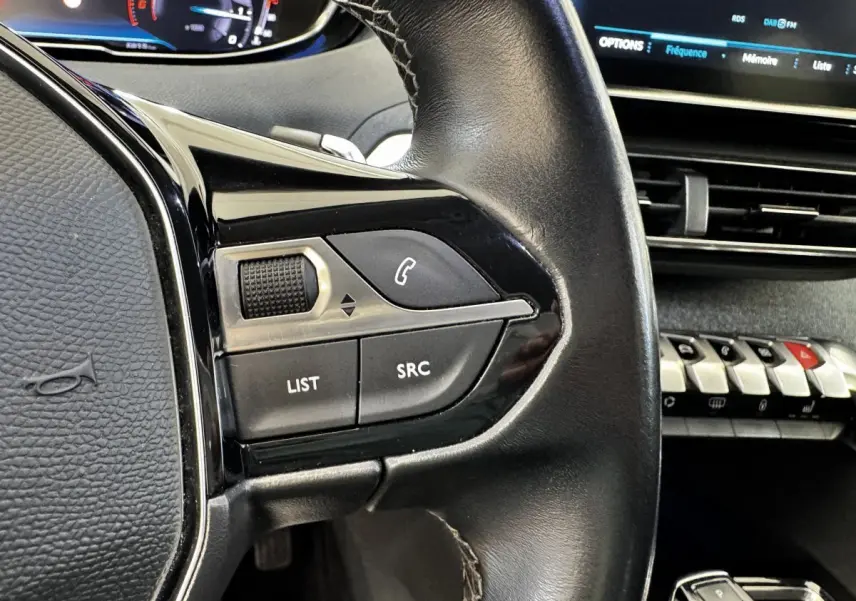 Gros plan sur le volant cuir noir de la Peugeot 5008 2022 avec commandes audio et tableau de bord numérique en arrière-plan.