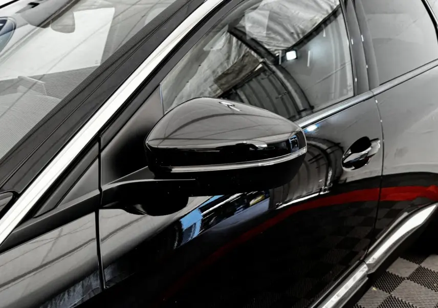 Gros plan sur le rétroviseur côté conducteur noir brillant d’un Peugeot 5008 noir, avec reflet de l’environnement intérieur.