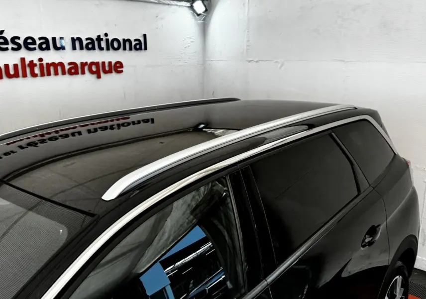 Vue 3/4 arrière droite du Peugeot 5008 noir, avec barres de toit chromées et vitres teintées dans un showroom.