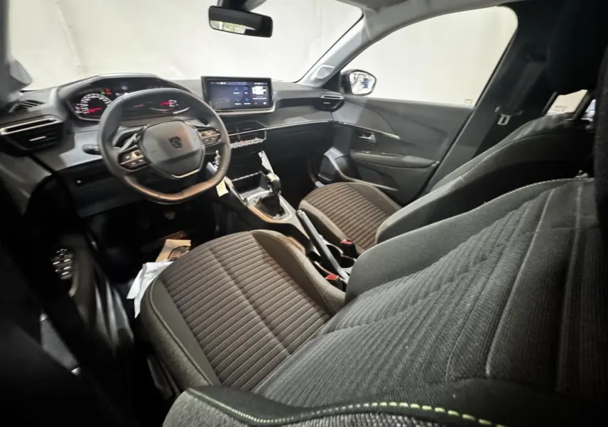 Vue intérieure côté conducteur de la Peugeot 208 2024, montrant le volant cuir, tableau de bord digital et sièges tissu gris.