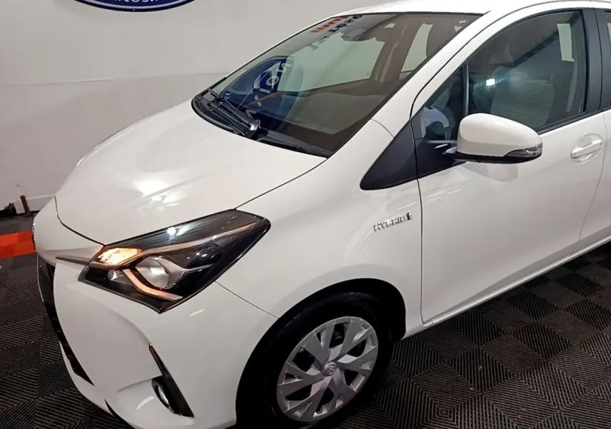 Toyota Yaris hybride blanche vue 3/4 avant droit, feux allumés et badge Hybrid visible sur l'aile avant.