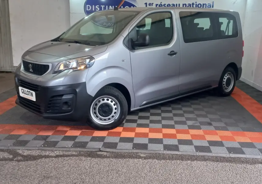 Vue 3/4 avant droit d’un Peugeot Traveller gris clair avec jantes acier et calandre noire, stationné en intérieur.