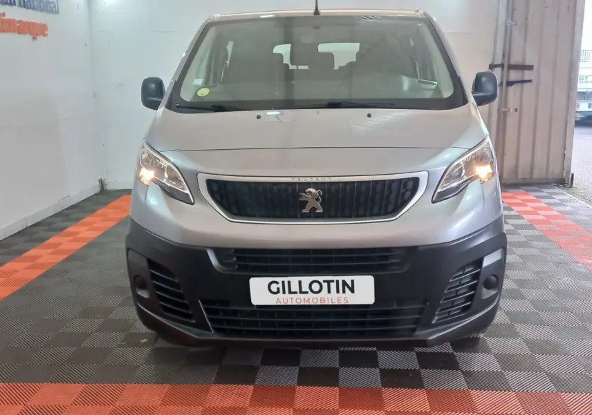 Vue frontale d’un Peugeot Traveller gris clair 2021 avec phares allumés et calandre noire, en intérieur showroom.