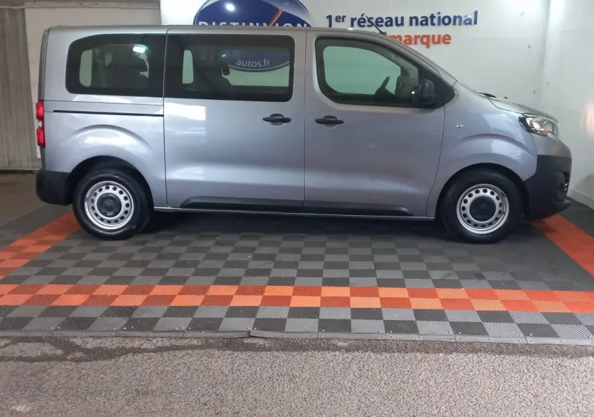 Vue de profil côté droit d’un Peugeot Traveller gris clair 2021 avec vitres arrière teintées et jantes acier simples.