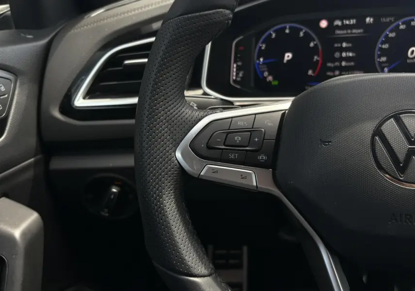 Vue rapprochée du volant cuir perforé côté gauche avec commandes régulateur adaptatif et tableau de bord digital du Volkswagen T-Roc noir intense 2025.