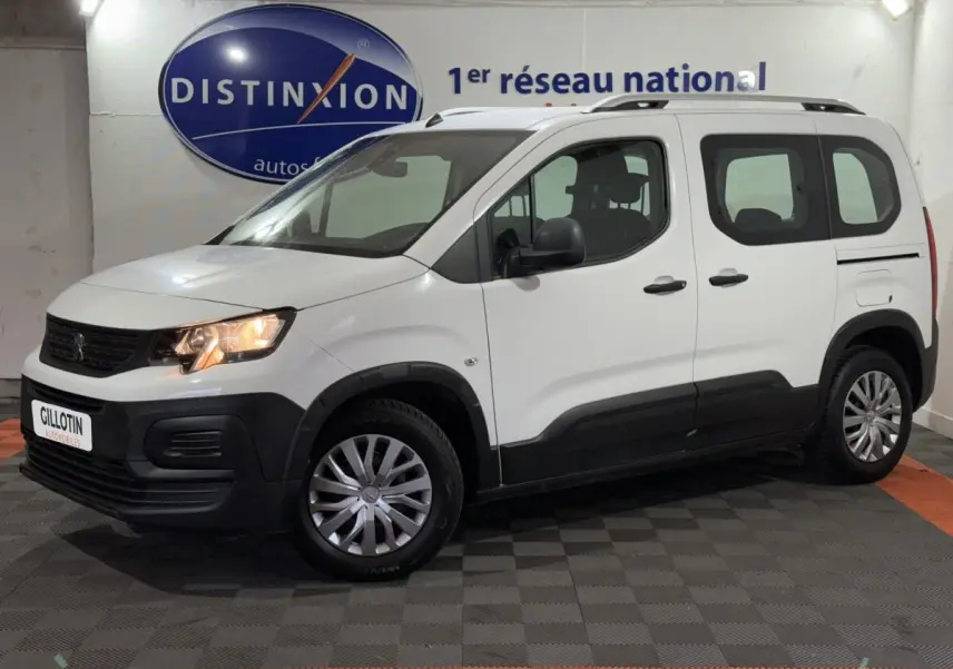 Peugeot Rifter blanc en vue 3/4 avant droit, avec barres de toit chromées et feux allumés dans un showroom.