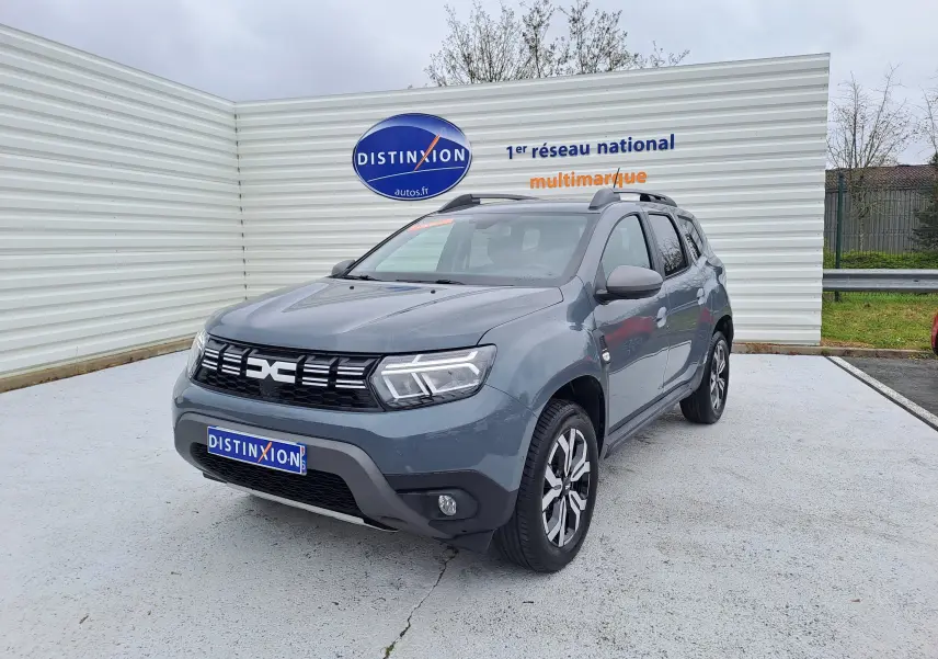 Dacia Duster gris urbain en 3/4 avant droit, avec jantes bi-ton et barres de toit visibles.