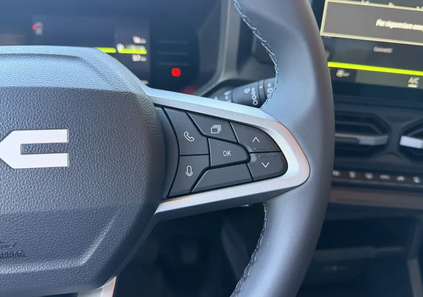 Gros plan sur le volant noir du Dacia Duster Hybrid 155 Journey 2026, avec commandes multifonctions à droite et tableau de bord numérique flou.
