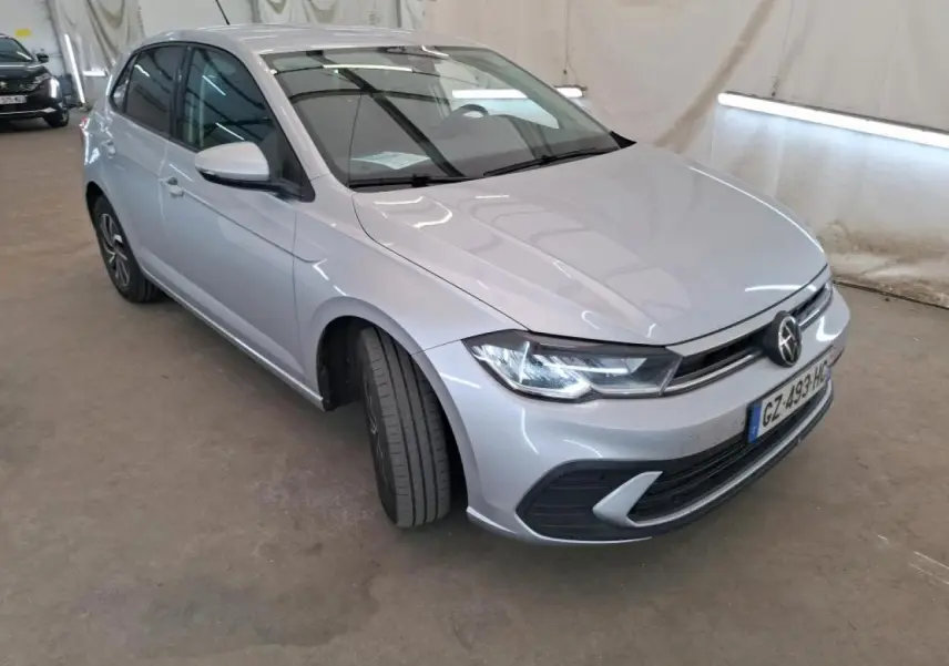 Volkswagen Polo gris clair vue 3/4 avant droit, avec calandre chromée et phares LED allumés.