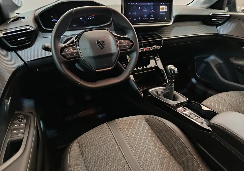 Vue intérieure côté conducteur de la Peugeot 208 PureTech 100 S&S Allure 2025, volant cuir et écran tactile central.
