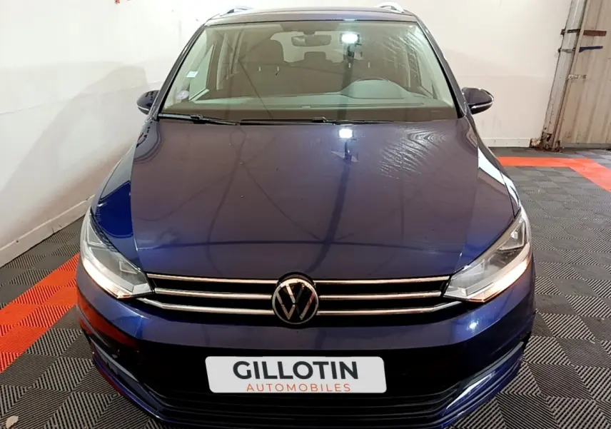Vue avant d'un Volkswagen Touran bleu 2021 avec calandre chromée et phares allumés dans un garage.