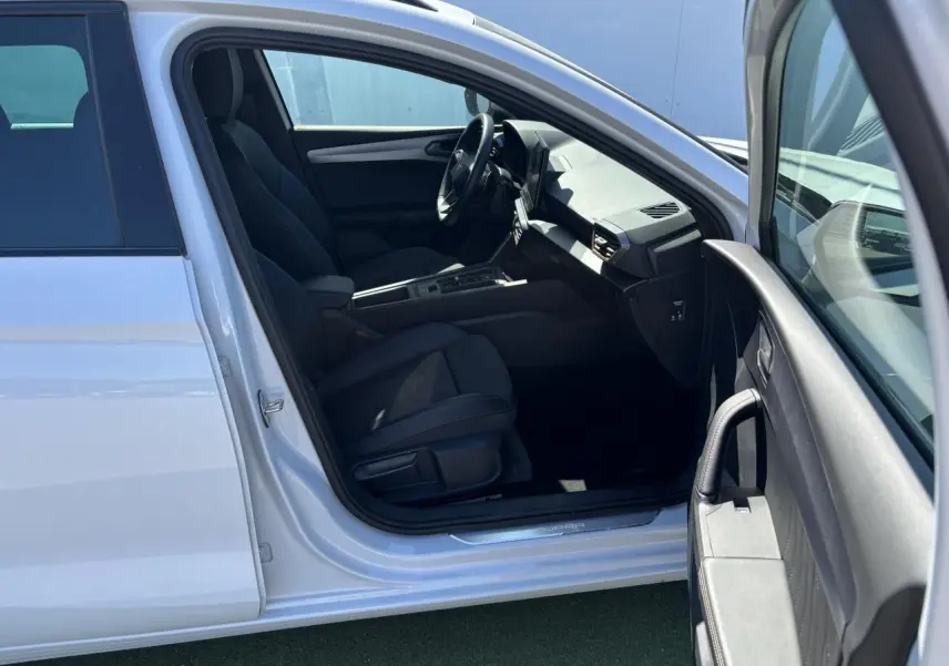 Vue intérieure côté conducteur d'une CUPRA Leon Sportstourer 2024 blanche Nevada, montrant sièges noirs et tableau de bord moderne.