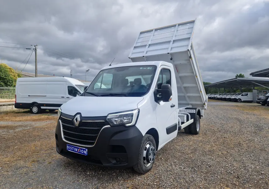 Renault Master Benne blanc en 3/4 avant droit, benne relevée et roues jumelées visibles sur terrain gravillonné.