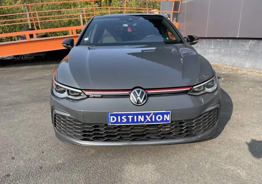 Vue de face d'une Volkswagen Golf VIII GTI gris métallisé avec calandre noire à motif nid d'abeille et bande rouge distinctive.