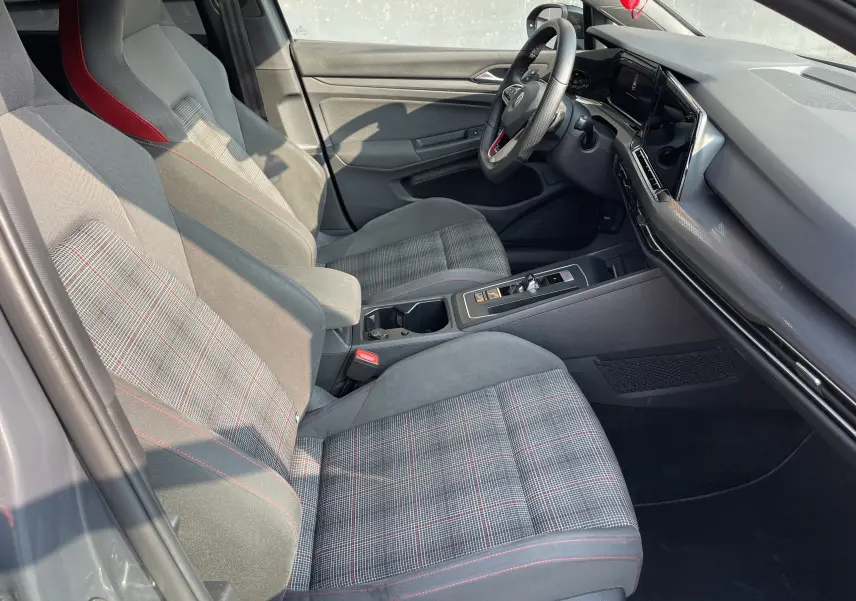 Intérieur côté conducteur de la Volkswagen Golf VIII GTI 2021, sièges tissu à motif écossais avec surpiqûres rouges et tableau de bord moderne.