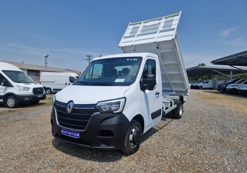Renault Master Benne blanc en 3/4 avant droit, benne relevée et roues jumelées visibles sur terrain gravillonné.