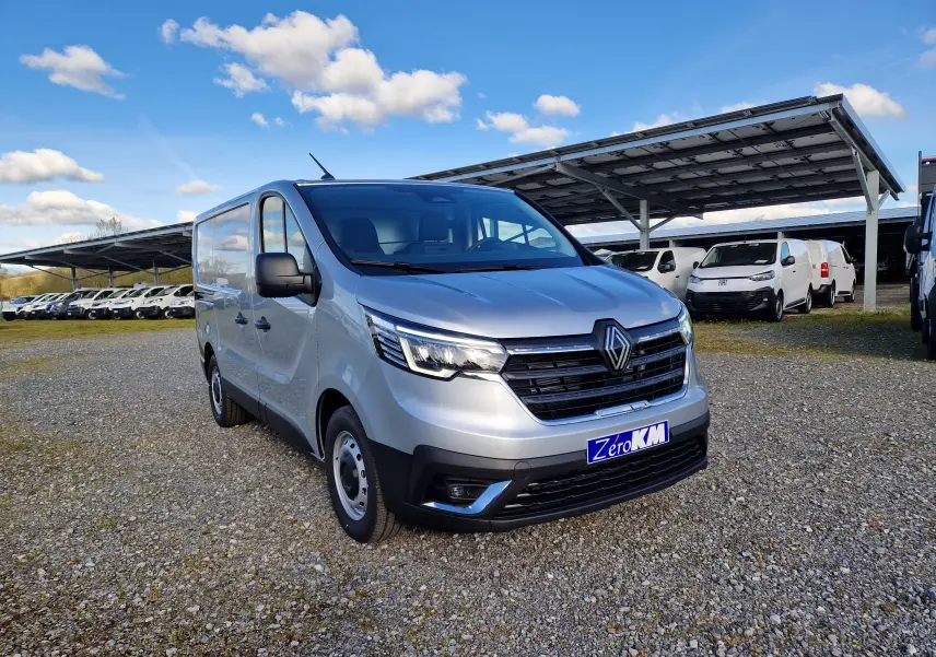 Renault Trafic Fourgon gris Highland vu en 3/4 avant droit sur parking avec plusieurs utilitaires en arrière-plan.
