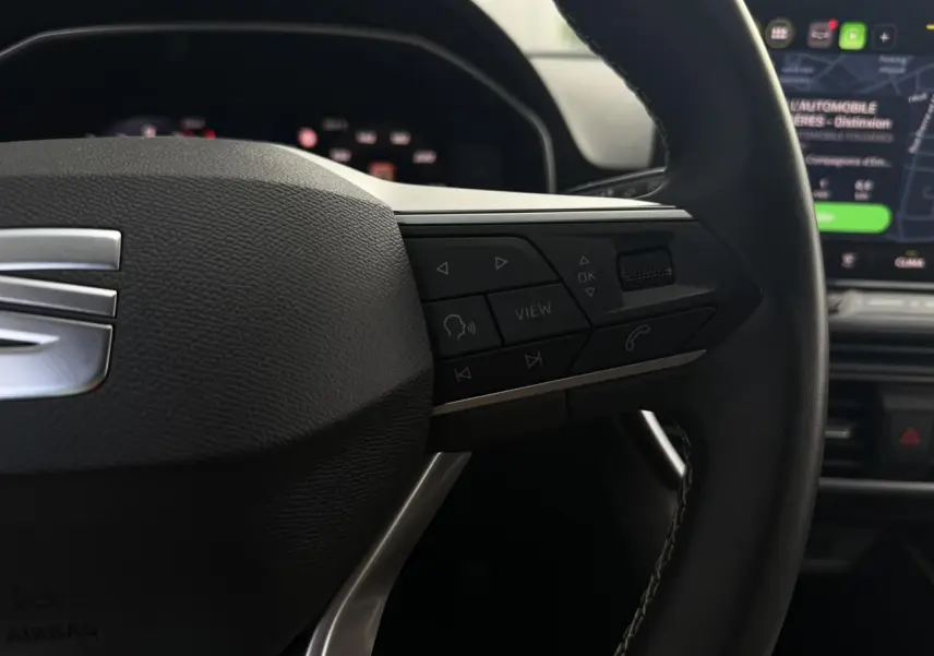 Gros plan sur le volant cuir multifonction noir du SEAT Leon ST 2025 avec commandes et écran tactile en arrière-plan flou.