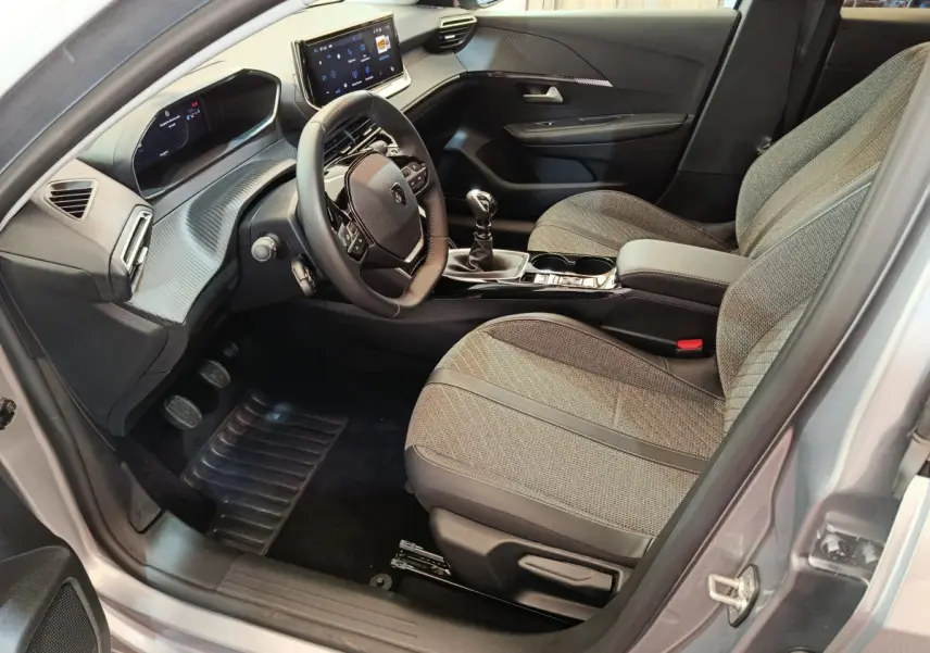 Intérieur avant droit du Peugeot 208 gris clair 2025, volant compact, écran tactile central et sièges tissu gris.