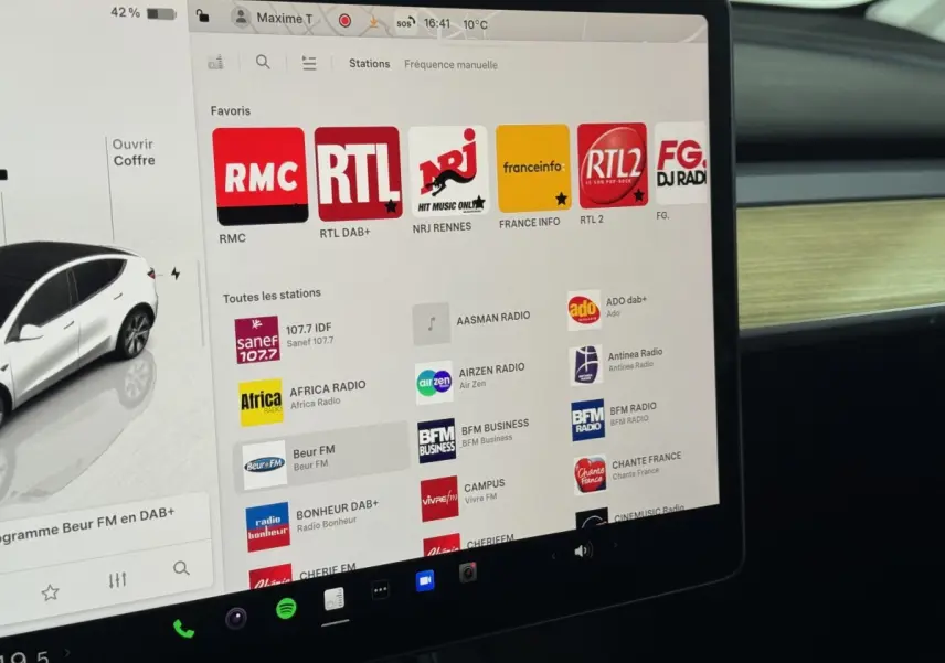 Écran tactile central de la Tesla Model Y blanc 2022 affichant les stations radio avec tableau de bord bois clair.