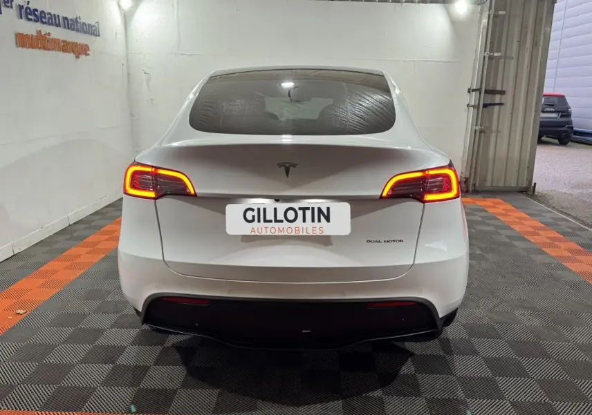 Vue arrière d'une Tesla Model Y blanche avec feux arrière allumés dans un garage éclairé.