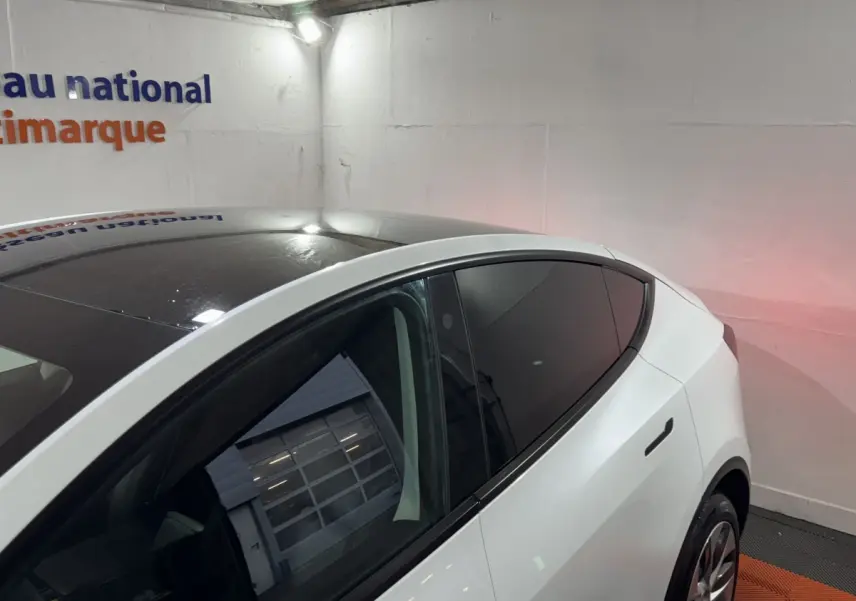Vue 3/4 arrière droite d'une Tesla Model Y blanche avec toit panoramique noir et vitres teintées dans un garage.