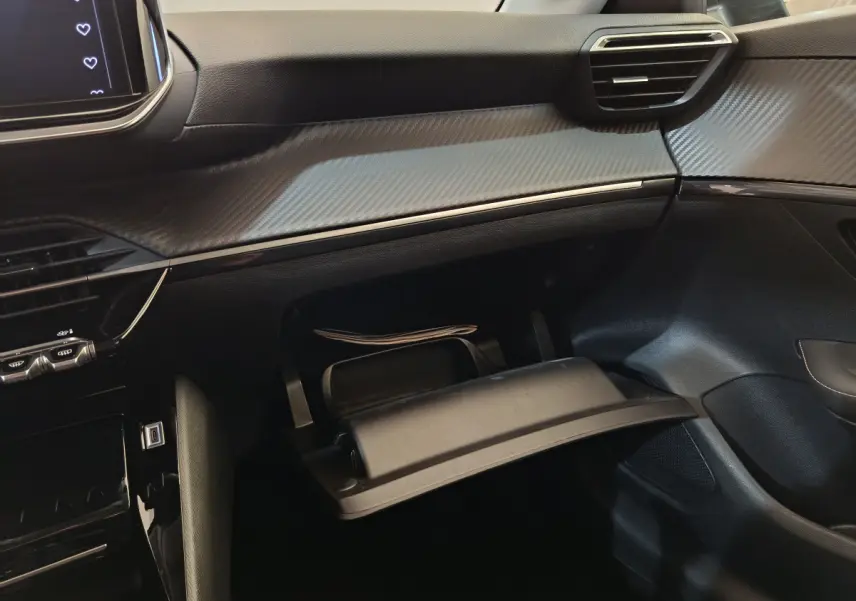 Vue intérieure côté passager de la Peugeot 208 gris clair 2025, montrant la boîte à gants ouverte et le tableau de bord avec finition carbone.