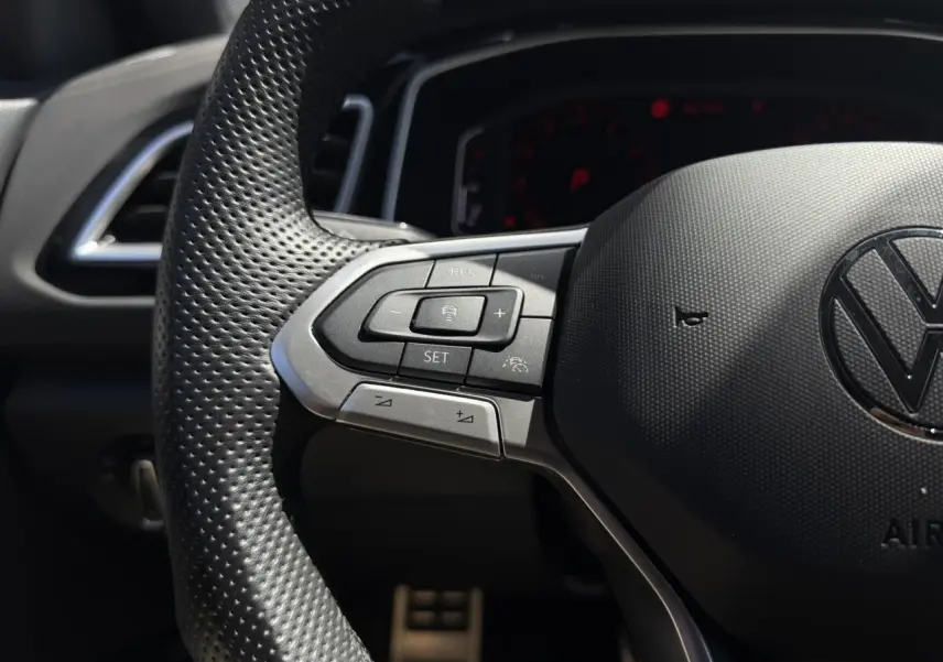 Gros plan sur le volant perforé noir du Volkswagen T-Roc 2025 avec commandes multifonctions côté gauche et logo VW central.