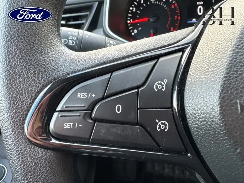 Gros plan sur les commandes du régulateur de vitesse au volant d'une Renault Clio blanche, tableau de bord en arrière-plan.