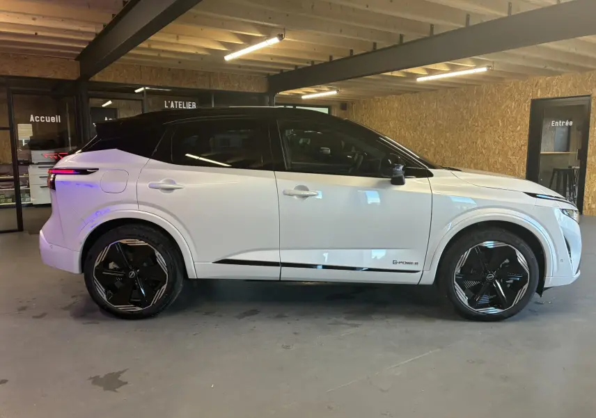 Profil côté droit du Nissan Qashqai 2025 blanc nacré avec toit noir et jantes noires en intérieur d'atelier.