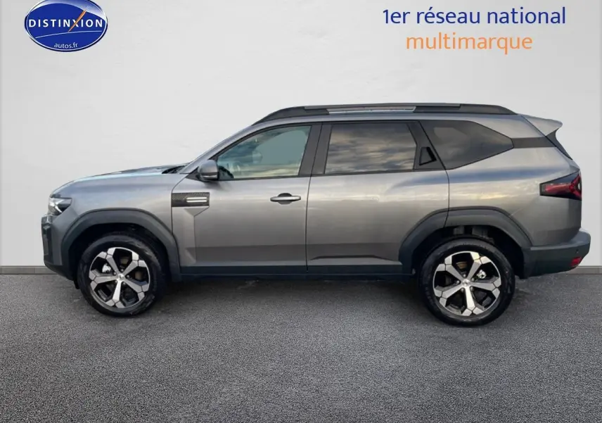 Vue de profil côté gauche du SUV Dacia Bigster gris schiste avec jantes alliage diamantées et barres de toit noires.